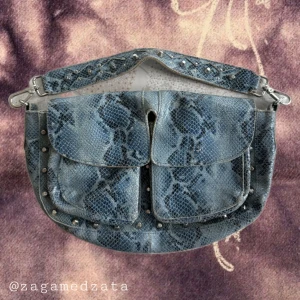 Blå axelväska i skinn med ormmönster - Snygg blå axelväska i skinn med ormmönster och silverfärgade nitar. Väskan har två stora fickor framtill, avtagbar axelrem och magnetlås. Invändigt finns dragkedjefack. Perfekt för dig som vill sticka ut med en unik accessoar.