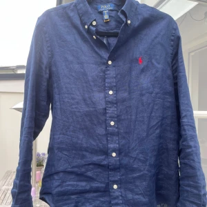 Marinblå linneskjorta Polo Ralph Lauren - Snygg marinblå skjorta från Polo Ralph Lauren i linne med klassisk button down-krage och vita knappar. Broderad röd logga på bröstet ger en stilren touch. Perfekt för dig som gillar en avslappnad men ändå uppklädd look. Från kidsbrandstore. Använd cirka tre gånger