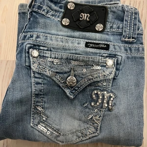 Miss Me ljusblå bootcut jeans med paljetter - Snygga ljusblå jeans från Miss Me med bootcut passform. Jeansen har detaljerade bakfickor med paljetter, broderier och silvriga nitar samt en broderad M-logga. Klassisk jeanslook med slitningar och dekorativa sömmar. Perfekta för dig som gillar statement-jeans. Längd: 102cn midja 36-37cm benöppning: 22cm inseam 82cm