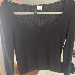 Svart ribbad topp med spets från Divided - Svart långärmad topp från Divided med ribbad struktur, söt spetskant runt den breda urringningen och en liten rosett framtill. Tight passform som sitter snyggt och funkar perfekt till jeans eller kjol.