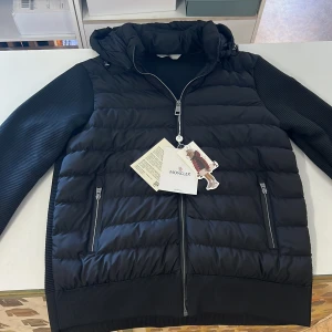 Svart cardiger från Moncler - Snygg svart pufferjacka från Moncler med ribbade ärmar och huva. Jackan har två dragkedjefickor framtill, hel dragkedja och ikonisk Moncler-logga på ärmen. Perfekt för dig som vill ha en stilren och exklusiv look under kyliga dagar. Finns i alla storlekar 