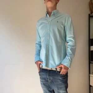 Ljusblå skjorta från Ralph Lauren - Snygg ljusblå skjorta från Ralph Lauren med klassisk button down-krage och broderad logga på bröstet. Skjortan har lång ärm, normal passform och stängs med vita knappar framtill. Perfekt för en clean och stilren look.