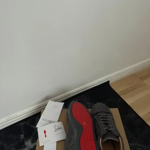 Gråa Louboutin  - Säljer ett par gråa sneakers från Christian Louboutin i mocka med ikonisk röd sula och coola nitar på tån. Skorna har snörning och en stilren, modern look som sticker ut. Perfekta för dig som vill ha något extra i din outfit.