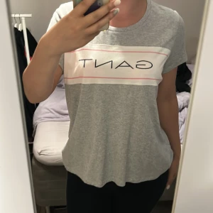 Grå GANT t-shirt med logga - Snygg grå t-shirt från GANT med klassisk logga tryckt över bröstet i svart. T-shirten har korta ärmar, rund halsringning och en vit panel med rosa linjer framtill. Skön och mjuk i bomull, perfekt till jeans eller shorts.