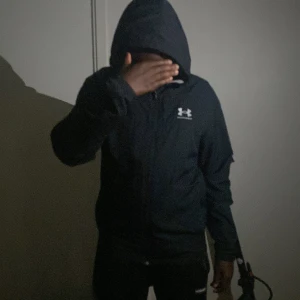 Svart vindjacka från Under Armour - Svart vindjacka från Under Armour med huva och dragkedja framtill. Jackan har en clean look med logga på bröstet och är tillverkad i lätt polyester. Perfekt för blåsiga dagar och enkel att matcha med streetwear. Snygg och funktionell för dig som gillar sportig stil.