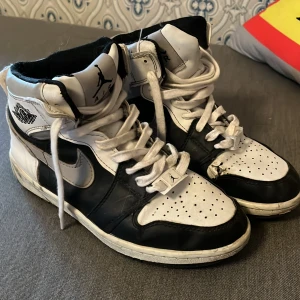 Nike Air Jordan 1 svartvita storlek 37  - Svartvita Nike Air Jordan 1 med klassisk high-top siluett, snörning och perforerad tå. Skorna har svart sula, vita och svarta paneler i skinn och ikoniska Jordan Wings-loggan på sidan. Perfekta för dig som gillar retrostil och streetwear.