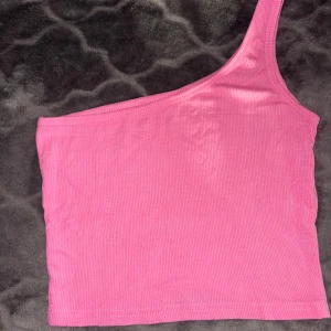 Rosa ribbad oneshoulder topp - Säljer en rosa ribbad topp med oneshoulder-design. Toppen är ärmlös och har en tight passform som framhäver axlarna. Perfekt för dig som vill sticka ut med en enkel men trendig look. Materialet är mjukt och stretchigt för extra komfort.