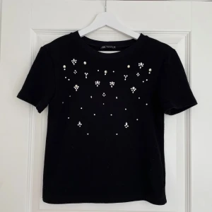 Svart topp med stenar från Zara - Svart kortärmad topp från Zara med rund hals och dekorerad med pärlor och små stenar framtill. Tjockare stickad känsla och rak passform. Perfekt för dig som vill ha något extra till din outfit. Inga defekter! Hör av er för frågor 💓