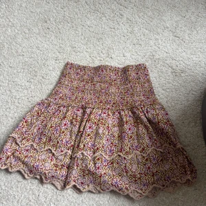 Blommig kortkjol från mango  - Supersöt kortkjol från mango med blommigt mönster i beige, rött och lila. Kjolen har smockad midja och volangig nederkant med vågig kant. Perfekt för sommarens alla äventyr och lätt att matcha med en enkel topp.