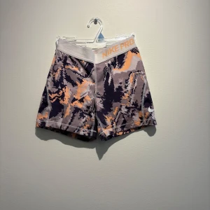 Nike Pro camo träningsshorts M - Nike Pro Dri-Fit shorts i camomönster med grå, svart och orangea toner. Tight passform och elastisk midja med Nike Pro-logga. Perfekta för träning och sportiga aktiviteter. Svalkande och snabbtorkande material.