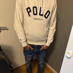 Vit hoodie från Polo Ralph Lauren - Snygg vit hoodie från Polo Ralph Lauren med stor broderad POLO-logga i mörkblått på bröstet och klassisk känguruficka. Tröjan har huva med snörning och långa ärmar med liten broderad logga vid ärmslutet. Perfekt för en chill och stilren look. Helt ny