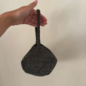 Svart glittrig kuvertväska med pärlor - Supersöt svart kuvertväska med glittriga pärlor över hela väskan. Den har en unik rundad form och ett smidigt handtag att bära runt handleden. 