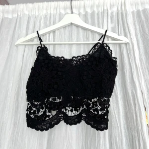 Svart topp - Supersnygg svart topp med tunna axelband och detaljerad spets med blommönster. Toppen har en vågig kant nertill och är perfekt för att lyfta vilken outfit som helst. Lätt och luftig känsla, perfekt för varma dagar eller när du vill sticka ut lite extra.
