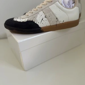 Maison Margiela sneakers med färgstänk - Maison Margiela sneakers i vitt och beige skinn med svarta färgstänk och beige mockadetaljer. Snygg kontrast mellan den vita ovandelen och den svarta tån, samt klassisk gummisula. Unik design med handmålat splash-mönster och diskreta logodetaljer på plösen.