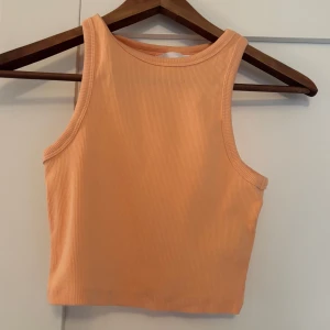 Orange ribbad linnetopp - Snygg croppad linnetopp i orange färg med ribbad struktur från HM. Ärmlös modell med hög halsringning, perfekt för varma dagar eller lager på lager. Enkel och stilren design som är lätt att matcha med jeans eller kjol.