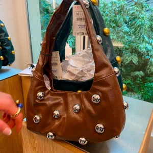 Brun axelväska med silvriga nitar - Cool brun axelväska i skinn med rundad form och stora silvriga nitar över hela väskan. Väskan har ett brett axelband och en trendig, chunky look som sticker ut. Perfekt accessoar för att lyfta vilken outfit som helst.