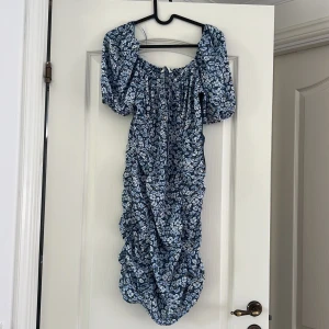 Blommig blå klänning med puffärm - Supersöt klänning i blått med vita och ljusblå blommor. Klänningen har puffiga korta ärmar med resår och smockad överdel med knytning framtill. Helt oanvänd med prislapp kvar 