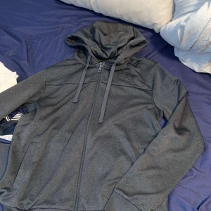 Grå hoodiejacka från EA7 Emporio Armani - Snygg grå hoodiejacka från EA7 Emporio Armani med dragkedja framtill och justerbar huva med snören. Jackan har diskret logga på bröstet och två fickor. Perfekt för en sportig och avslappnad stil.