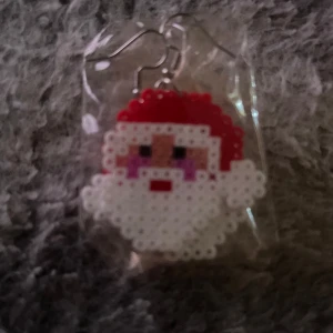 Juliga örhängen med pärlplatta tomte - Unika örhängen med en söt tomte gjord av pärlplattor. Perfekt accessoar för att sprida julkänsla! Färgerna är röd, vit, rosa och svart. Hängsmycket har en pixelart-look och är riktigt charmigt.