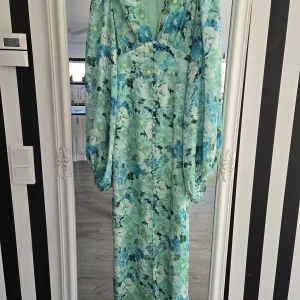 Adoore Positano långklänning mintblommig - Superfin långklänning från Adoore i modellen Positano. Klänningen har ett mintgrönt blommigt mönster, v-ringning, knappar framtill och vida långa ärmar. Perfekt för dig som vill sticka ut med en färgstark och romantisk look.