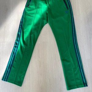 Gröna Adidas mjukisbyxor med blå ränder - Snygga gröna mjukisbyxor från Adidas med klassiska blå ränder längs sidorna och dragkedjor vid fickorna. Byxorna har rak passform och elastisk midja för skön komfort. Perfekta för chill eller träning.