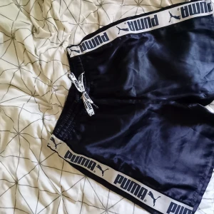 Svarta Puma shorts med logoband - Marinblå shorts från Puma med breda vita logoband längs sidorna och midjan. De har elastisk midja med snörning och är tillverkade i glansig polyester. Perfekta för sport eller chill, klassisk loose fit och meshfoder för extra komfort.