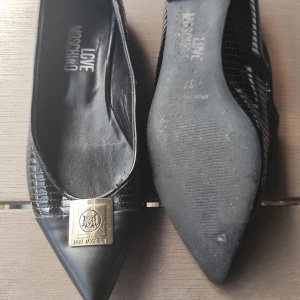 Svarta ballerinaskor Love Moschino - Snygga svarta ballerinaskor från Love Moschino med spetsig tå och guldfärgad metallplatta framtill. Skorna har en elegant struktur i skinnimitation och en klassisk, stilren look som passar till många outfits.