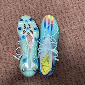 Adidas X Speedportal blå fotbollsskor - Säljer ett par Adidas X Speedportal fotbollsskor i ljusblått med regnbågsfärgade ränder och coola detaljer. Skorna har snörning, platt sula med dobbar och inslag av gult och rött. Perfekta för dig som vill sticka ut på planen! Helt nya elite Skor