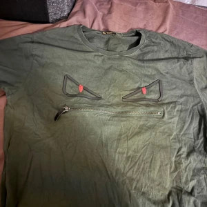 Grön Fendi t-shirt med dragkedja - Säljer en grön t-shirt från Fendi med coola ögon-detaljer och en dragkedja framtill som öppnar upp till ett svart innerfoder med vita tryckta ord. T-shirten har rund halsringning och korta ärmar. Perfekt för dig som vill sticka ut med en unik design!