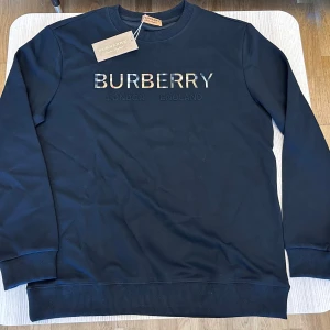 Svart sweatshirt från Burberry - Snygg svart sweatshirt från Burberry med klassisk logga broderad på bröstet. Tröjan har rund hals, långa ärmar och ribbade muddar. Perfekt för dig som gillar stilrena och exklusiva plagg med streetkänsla.