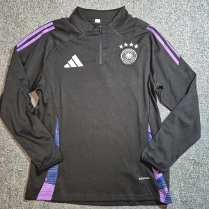 Svart långärmad Tyskland Adidas tröja - Snygg svart långärmad fotbollströja från Adidas med Tysklands landslagsemblem och fyra stjärnor på bröstet. Tröjan har lila detaljer på axlar och sidor, halv dragkedja och är tillverkad i funktionsmaterial Aeroready. Perfekt för träning eller match.