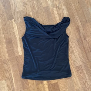 Svart topp från BikBok - Snygg svart topp från BikBok i stretchigt och glansigt material. Toppen har en draperad axel och sitter tight mot kroppen, perfekt för fest. Enkel men ändå unik design som passar till många olika looks. Helt oanvänd 💞