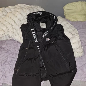 Svart dunväst från Moncler - Svart dunväst från Moncler med stor huva och tydlig logga på bröstet. Västen har dragkedjor på fickorna och längs framsidan samt breda snören med Moncler-tryck. Snygg, puffig design som ger en sportig vibe och passar perfekt till lager-på-lager-stil.