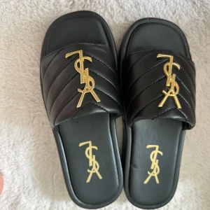 Svarta sandaler med guld YSL-detalj - Svarta sandaler från YSL med quiltad ovandel i skinn och ikonisk guldig YSL-logga framtill. Platt sula och öppen tå ger en clean och lyxig vibe. Perfekta för att lyfta din outfit med en exklusiv touch. Oanvända, inspirerade 