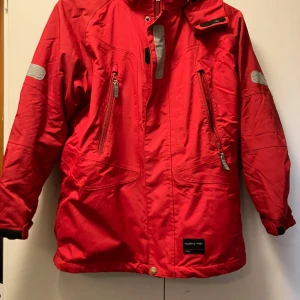 Röd vinterjacka Polarn O. Pyret 146 - Röd vinterjacka från Polarn O. Pyret med svarta detaljer och reflexer på ärmarna. Jackan har dragkedjor framtill, hög krage och praktiska fickor. Insidan är fodrad i svart och har snölås med knappar. Perfekt för kalla dagar och snygg stil. Hel och ren! Jag kan inte hitta något att anmärka på.