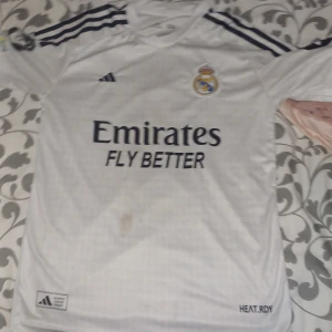 Real Madrid Mbappé 9 fotbollströja Adidas - Säljer en vit Real Madrid fotbollströja med Mbappé och nummer 9 på ryggen. Tröjan har korta ärmar, svarta Adidas-ränder på axlarna och klubbmärke på bröstet. Materialet är lätt och andas, perfekt för match eller träning. Emirates Fly Better tryck på framsidan.