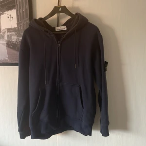 Mörkblå hoodie från Stone Island - Snygg mörkblå hoodie från Stone Island med dragkedja, huva och klassisk logga på ärmen. Tröjan har känguruficka framtill och justerbara snören i huvan. Perfekt för dig som gillar märket Stone Island. 