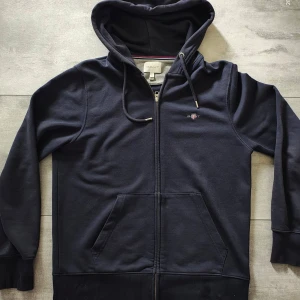 Mörkblå hoodie från GANT med dragkedja - Mörkblå hoodie från GANT med hel dragkedja, huva med snörning och broderad logga på bröstet. Tröjan har känguruficka framtill och ribbade muddar vid ärmslut och nederkant. Perfekt för chill dagar eller när du vill ha en clean look. (Knappt använt)
