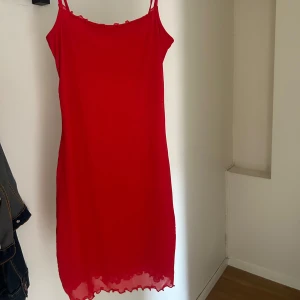 Röd ärmlös kortklänning med volangkant - Snygg röd kortklänning med tunna axelband och volangkant nertill. Klänningen har en tajt passform men är väldigt stretchigt i materialet. Materialet känns lätt och luftigt, vilket gör den skön att bära under varma dagar.