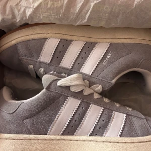 Grå Adidas Campus sneakers - Snygga grå Adidas Campus sneakers i mocka med de klassiska tre vita ränderna på sidan. Skorna har en vit sula och snörning framtill. Sparsamt använda och är vi snyggt skick! 