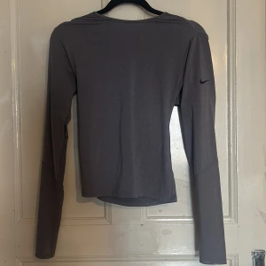 Grå långärmad topp från Nike - Snygg grå långärmad topp från Nike med omlott-design framtill. Perfekt för en stilren och sportig look. Den har en diskret Nike-logga på ärmen.