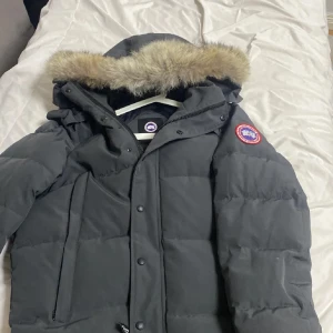 Svart dunjacka med päls från Canada Goose - Säljer en svart dunjacka från Canada Goose med en mysig pälsfodrad huva. Jackan har en klassisk design med dragkedja och knappar framtill samt en logotyp på ärmen. Perfekt för kalla vinterdagar!