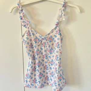 Linne - Jätte söt blommit linne i vitt med blå och rosa. Toppen har volangdetaljer vid axelbanden och en liten rosett framtill. Perfekt för varma sommardagar!