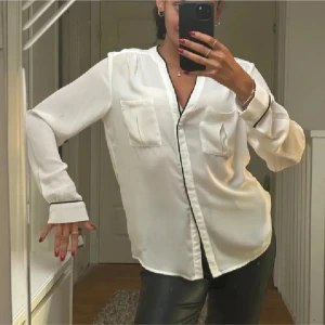 Vit blus med svarta detaljer - Snygg vit blus med svarta kantdetaljer och två bröstfickor. Blusen har långa ärmar och en avslappnad passform, perfekt för en stilren look.