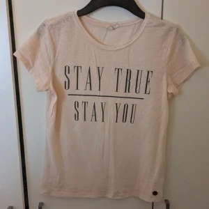 Beige t-shirt med tryck - Säljer en beige t-shirt med texten 'STAY TRUE STAY YOU' tryckt på framsidan. T-shirten har en rund hals och korta ärmar. Perfekt för en avslappnad stil.