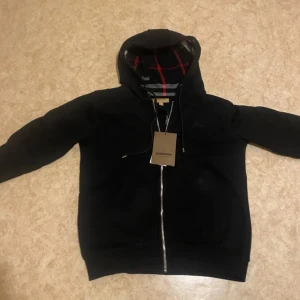 Svart hoodie med dragkedja från Burberry - Snygg svart hoodie från Burberry med dragkedja och huva. Insidan av huvan har ett klassiskt Burberry-rutigt mönster i rött, vitt och svart. Perfekt för en stilren look.