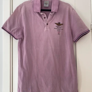 Lila pikétröja från Aeronautica Militare - Säljer en snygg lila pikétröja från Aeronautica Militare med broderad logga på bröstet. Tröjan har korta ärmar med randiga muddar och en klassisk krage med knappar. Perfekt för en stilren look!  Lila tvättad piké i strl M från Aeronautica Militare.  Använd men i fint begagnat skick.  Mått Över bröstet: ca 51 cm Längd från axeln och ner: ca 71 cm  100% bomull  Kommer från ett djur och rökfritt hem. 