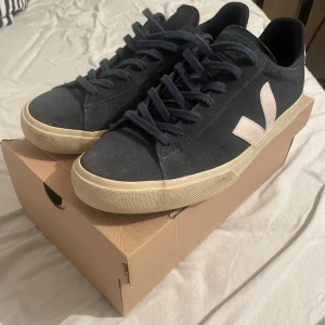 Marinblåa sneakers från Veja - Snygga marinblåa sneakers från Veja med vita detaljer och snörning. Skorna har en klassisk design med en bekväm sula och är perfekta för vardagsbruk. De är tillverkade i mocka och har en stilren look. Tveka inte på att lägga ett bud😃