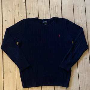 Navy Ralphlauren cableknit - Säljer en stilren mörkblå stickad tröja från Ralph Lauren med kabelstickat mönster. Tröjan har långa ärmar och en liten röd logotyp på bröstet. Perfekt för kyligare dagar! Tröjan passar till XL barn.