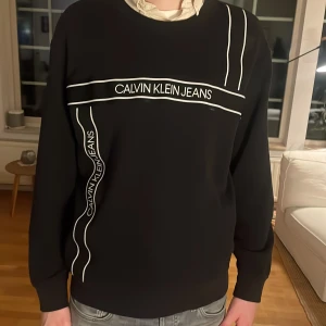 Svart tröja från Calvin Klein Jeans - Snygg svart tröja från Calvin Klein Jeans med vita linjer och logotyp på framsidan. Tröjan har långa ärmar och en klassisk passform. Perfekt för en stilren look.